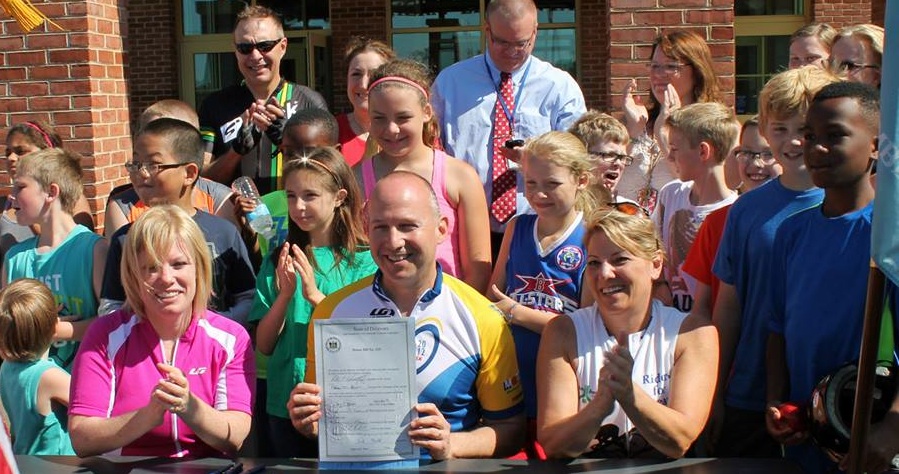 Governor Jack Markell (center) shows off the bill making cycling the Official Sport of Delaware while bill sponsors Senator Nicole Poore (left) and Representative Valerie Longhurst (right) and kids from Wilbur and Southern Elementary Schools applaud.
